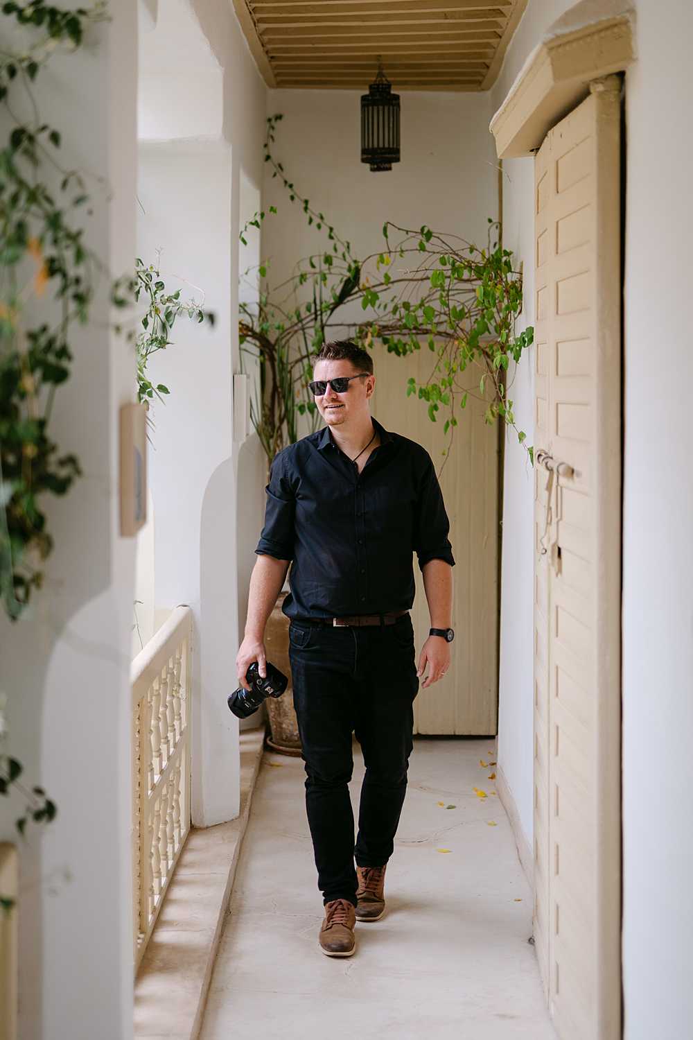 Ross Hurley photographing a wedding in Morocco, walking through a white riad-style corridor with his camera.