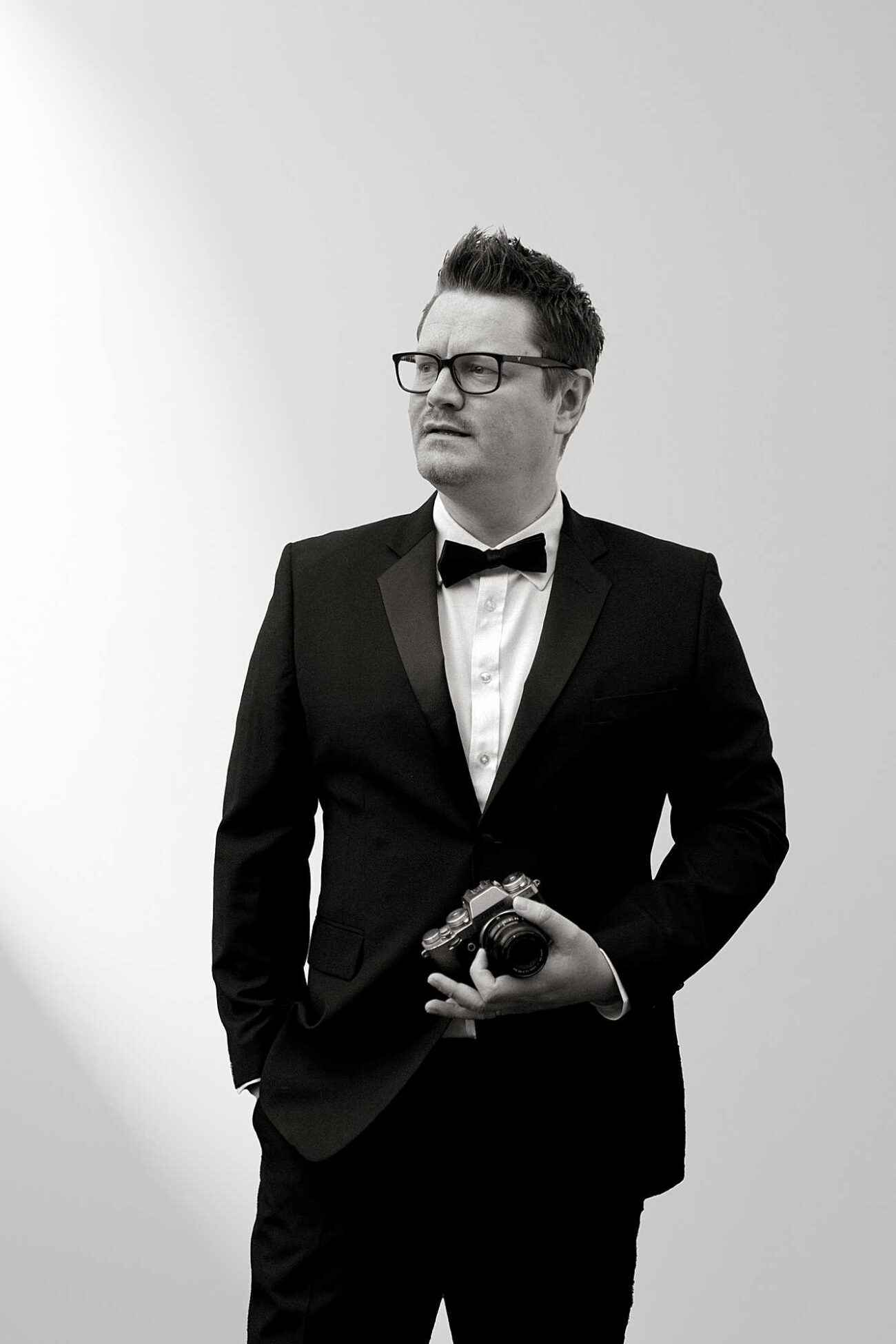 Ross Hurley, UK and destination wedding photographer, in a tuxedo holding a camera.
